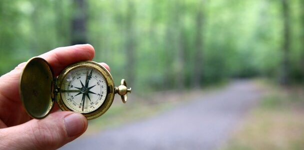 Two fingers holding a compass in front of a forest trail.